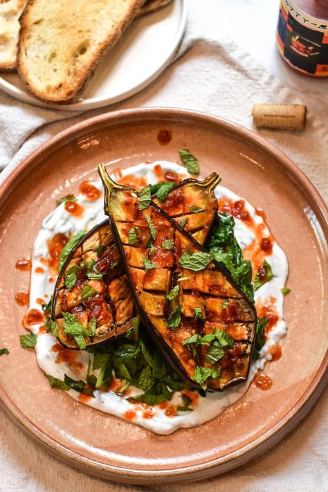 A terracotta colored platter with a spread of yogurt, a spinach herb salad, two cross hatched roasted eggplants, drizzle of harissa oil, and fresh chopped herbs. Partially out of frame is a plate of sourdough toast, a bottle of wine, and a wine cork. Eggplant Salad Recipes Healthy, Eggplant Yogurt Recipe, Spicy Garlic Eggplant, Halloumi Eggplant, Harissa Aubergine, Harissa Eggplant, Yacht Food, Eggplant Harissa, Roast Aubergine