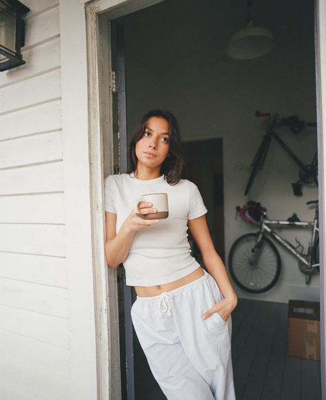 A woman with a cup of coffee leans against the door frame to outside in pajamas Lazy Day Photoshoot, Door Lean Pose, Door Frame Poses, Leaning Against Door Frame Pose, Pose Berdiri, Leaning Pose, Coffee Pose, Skincare Shoot, Woman Lounging