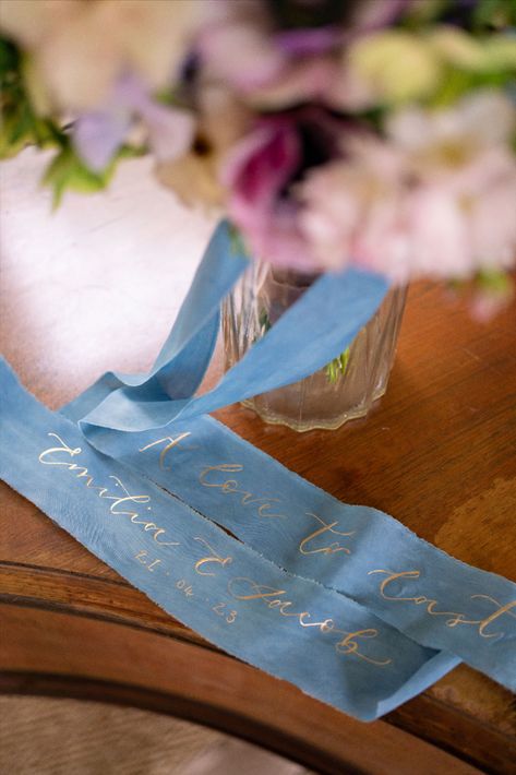 At the top there is a floral wreath out of focus, below lay two dusty blue ends of a bouquet ribbon with the words 'a love to last' and the couples name and their wedding date below hand foiled in gold. Quote In Calligraphy, Gold Foil Stationery, Calligraphy Photography, Wedding Bouquet Ribbon, Calligraphy A, Bouquet Ribbon, Silk Wedding Bouquets, Silk Bouquet, Ribbon Bouquet