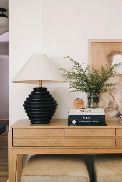 Recreate this styled console table by sticking with neutral colors and natural materials keep. The black and white colors of the chic table lamp are repeated in a stack of books next to it, a glass vase with greenery livens up the surface, and a leaning piece of art grounds the neutral decor. #consoletableideas #entrywayideas #entrywaytableideas #bhg Glass Vase With Greenery, Black Console Table Decor, Console Table Decor Ideas, Vase With Greenery, Style A Console Table, How To Style A Console Table, Layered Artwork, Chic Table Lamp, Console Table Decor