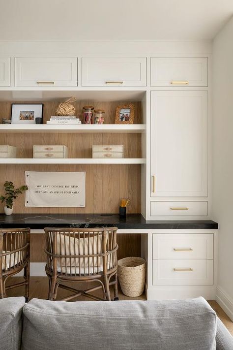 Family room features a black marble top built in desk with brown bamboo chairs under shelves surrounded by white cabinets with brass pulls,