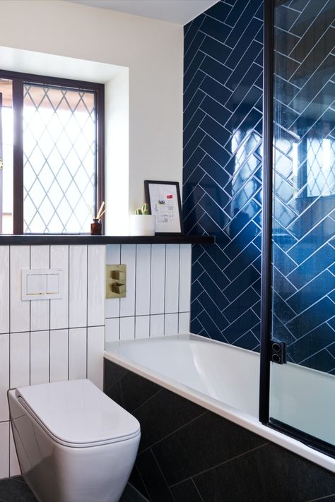 A modern bathroom with a striking navy blue herringbone tile wall surrounding the bathtub, complemented by a black-framed glass shower screen. Adjacent to the bath, a vertical white tile wall adds contrast and sophistication, adorned with a brushed brass flush plate and fixtures. The overall design is sleek, stylish, and contemporary with a bold colour palette and refined details. Small Family Bathroom, Family Bathroom Design, Unique Bathrooms, Herringbone Tiles, Chevron Tile, Traditional Bathrooms, Chic Bathroom, Cage Light, Kitchen Company