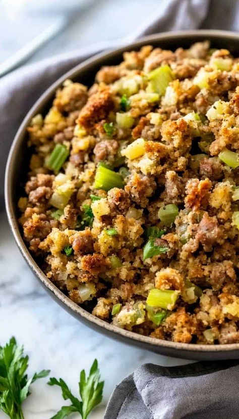 A close-up shot of a bowl filled with golden brown sausage stuffing, featuring chunks of Italian sausage, celery, and bread cubes. The dish is garnished with fresh parsley and set against a light-colored marble surface with a grey napkin beside it. Thanksgiving Casserole Recipes, Green Bean Casserole Crock Pot, Bacon Wrapped Green Beans, Creamy Mashed Cauliflower, Thanksgiving Stuffing Recipes, Cherry Tomato Salad, Pumpkin Mac And Cheese, Honey Roasted Carrots, Sausage Stuffing