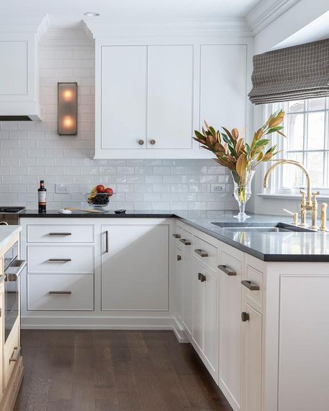 White inset kitchen cabinets adorned with brass pulls and a black quartz countertop are fitted with a sink matched with an antique brass deck mount faucet fixed beneath a window framed by white glass subway backsplash tiles. White Cabinets Black Countertops, White Transitional Kitchen, New Build Interior, Kitchen Space Ideas, Modern Traditional Kitchen, Black Quartz Countertops, White Cabinets White Countertops, Kitchen Hood Ideas, Tiny Kitchen Remodel