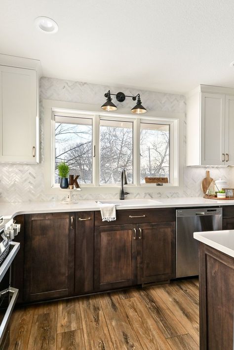 The kitchen features two-toned cabinets - stained alder for the island/lowers and off-white upper cabinets. A marble herringbone backsplash compliments the space with a mix of cool/warm tones. Angled plug molding and undercabinet lighting keep the backsplash uninterrupted. Adding to the beauty is the satin bronze hardware and a custom hood with a wood shelf detail. White Upper Cabinets Dark Lower Backsplash, Brown Base Cabinets White Upper, Umber Cabinets Kitchens, Wood Cabinets Chrome Hardware, Brown Cabinets Silver Hardware, Wood Trim White Cabinets, Kitchen With Few Upper Cabinets, Stained Bottom Cabinets Painted Top, Wooden Lower Cabinets White Uppers