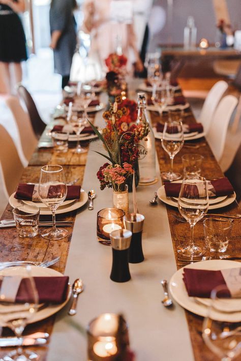 Schlichte Dekoidee für eine lange Hochzeitstafel mit rustikalen Holztischen: In der Mitte verläuft ein neutraler Tischläufer in beige. Darauf stehen kleine Windlichter, sowie kleine Vasen mit dezenten Blumenarrangements. Diese passen mit ihren Rottönen zu den bordeauxroten Servietten. Weiße Teller und Silber-Besteck runden die Hochzeits-Tischdeko ab. → Gesehen bei einer Hochzeit in der Location Holsteins Mühle auf djmarkusrosenbaum.de | Tischdeko Hochzeit schlicht | Foto: Tim Kurth Quick Saves