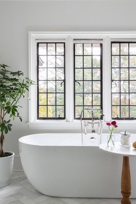 An oval freestanding bathtub with a chrome vintage tub filler sits on marble herringbone backsplash tiles beneath black framed windows. Black Framed Windows, Dark Stained Wood Floors, Marble Pedestal Dining Table, Painted Wood Chairs, Oval Bathtub, Herringbone Tile Backsplash, Framed Windows, Tub Design, Marble Herringbone