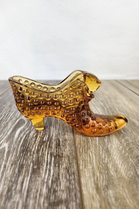 This Fenton Art Glass amber boot is a stunning piece of vintage décor. Crafted with Fenton's signature hobnail pattern, this glass shoe adds a touch of elegance to any collection. Perfect as a unique gift or to display on a shelf, its rich amber hue and intricate design make it a timeless treasure for glassware enthusiasts. Glass Shoe, Glass Shoes, Glass Gifts, Timeless Treasures, Slipper Boots, Amber Glass, Vintage Shoes, Vintage Decor, Art Glass