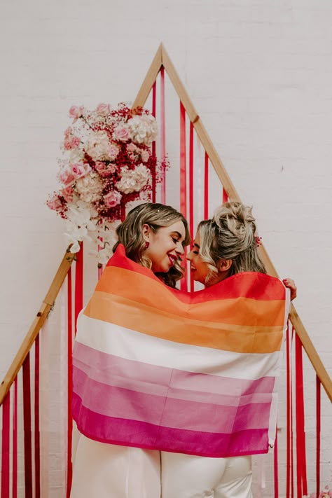 Brides wrapping themselves up in lesbian flag standing in front of wooden triangular wedding arch with pink and red ribbons hanging from it and pink rose, peony, carnation and dried flower decor | Gabriela's Photography And Film Lesbian Wedding Ideas Decor, Red Orange Yellow Pink Wedding, Pride Wedding Ideas, Lesbian Flag Decor, Lesbian Flag Wedding, Lesbian Cake Topper Wedding, Lesbian Wedding Suit, Lesbian Wedding Outfits, Lesbian Destination Wedding