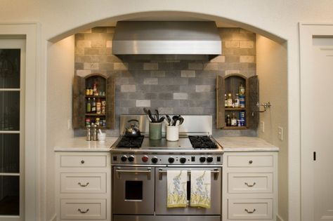 Arched cooking alcove with stainless steel stove framed by ivory drawers below a white marble countertop with a pair of arched mini spice cabinets hung over a gray brick style backsplash with stainless steel vent hood and side wall mounted pot filler faucet. Traditional Colonial Interior, Cooking Alcove, Gray Tile Backsplash, Blue Mosaic Tile, Stainless Steel Stove, Colonial Interior, Traditional Colonial, Cottage Kitchens, White Marble Countertops