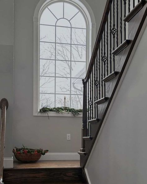 Dark wooden steps let down to a landing that features soft gray walls and a gorgeous arched window styled for the holidays with a garland and simple candle holder. The trim of the window is a classic white that matches the grilles. Modern Stairway, Stair Bookshelf, Staircase Window, Modern Staircase Design, Stairway Wall, Staircase Design Ideas, Bookshelf Modern, Stairs Window, Simple Candle