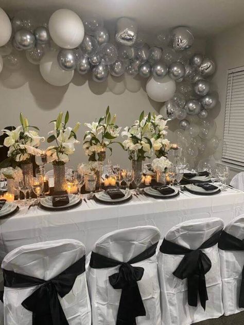 Glam up the dinner table for your family reunion with a charming white tablecloth and top it with a black runner. Line up tall cylindrical glass vases adorned with rhinestone wraps along the center and insert faux calla lily stems in them. Place white dinner plates, cutlery, and black linen napkins to perfect the setup while decorating the backdrop wall with white, silver latex, and orbz balloons. Also, cover the chairs with polyester white covers and black sashes for fabulous grandeur. Event Decor Ideas, Party Decorations Table, Backdrop Wall, White Tablecloth, Black Runners, White Dinner, White Dinner Plates, White Table Cloth, Glass Vases