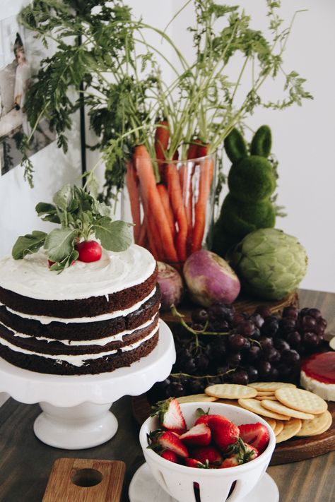 Fresh produce is arranged on a table, alongside a layered chocolate cake with radishes atop. Fall Peter Rabbit Party, Beatrix Potter First Birthday, Peter Rabbit Food Ideas, Peter Rabbit Tablescape, Peter Rabbit First Birthday Girl, Vintage Peter Rabbit Baby Shower Ideas, Peter Rabbit Party Food, Peter Rabbit 1st Birthday Boy, Rabbit Birthday Theme