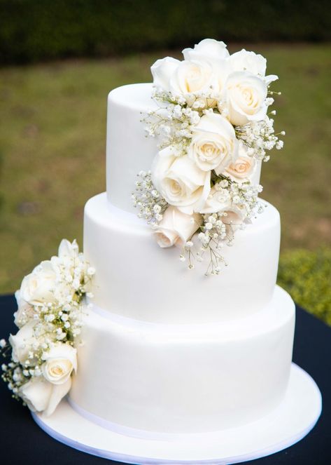 A White and beautiful wedding cake. The white wedding cake is decorated with white roses. The white wedding cake is made into three tiers. Simple Three Tier Wedding Cake With Flowers, Plain White 3 Tier Wedding Cake, Wedding Cakes With Gypsophilia, White Roses On Wedding Cake, Three Tier White Wedding Cake, Wedding Cake With Baby Breath Flowers, 3 Tier White Wedding Cake With Flowers, White Wedding Cake With White Flowers, Wedding Cake With Pink Roses