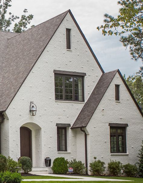 home exterior + lantern sconce + landscaping + greenery + pointed roof + gray roofing + white brick + dark gray windows + arched doorway | greg busch Roof Color With White House, Dark Roof White House, Brick Dormer, Cream Brick House, White House Brown Roof, Country Cottage Plans, Landscaping Greenery, Gray Windows, Painted Brick Exteriors