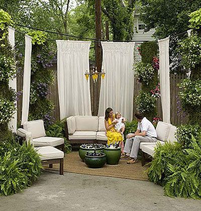 Tall white drapes are hung around the driveway space with a loveseat with cotton cushions and side chairs arranged in a u-shape to create an outdoor room. Ferns in containers and other hanging flowers are hung throughout the space. Yard Privacy, Patio Privacy, Yard Party, Backyard Privacy, Outdoor Privacy, Outdoor Curtains, Outdoor Landscaping, Porch Patio, Outdoor Rooms