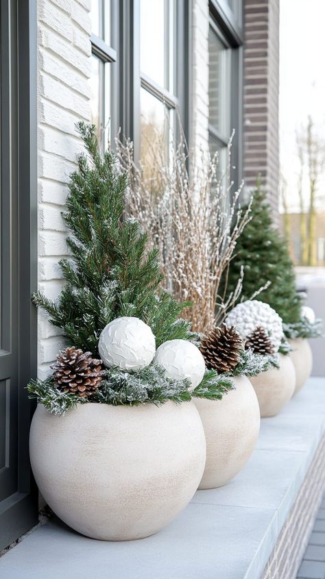 Five large beige planters with pine cones, evergreen branches, and white Christmas balls on a Dutch house's winter porch. Winter Porch Planters, Outdoor Pots For Christmas, Outdoor Christmas Planters Winter Porch Front Doors, Winter Greenery Outdoor Pots, Diy Winter Planter Front Porches, Winter Planter Ideas Outdoor, Christmas Porch Planter Ideas, After Christmas Winter Decor Porch, Christmas Outdoor Pots Planters