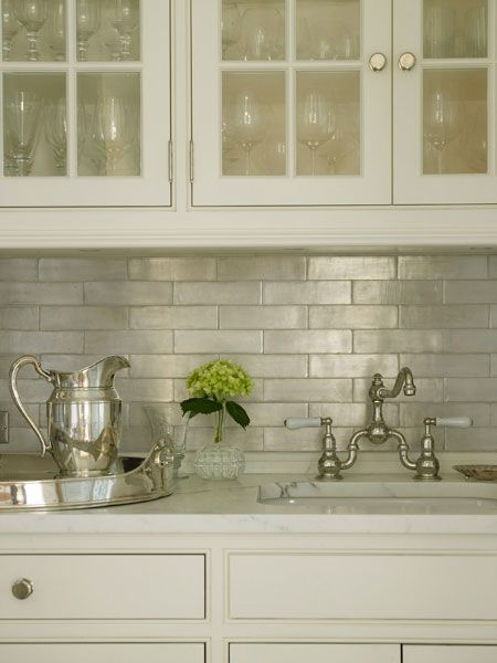 Glass-front upper cabinets and ivory lower cabinets paired with white marble countertops and iridescent tile backsplash laid out in brick pattern framing sink with vintage style bridge faucet. Ivory Cabinets, Glass Backsplash Kitchen, Iridescent Tile, Farmhouse Backsplash, Brick Backsplash, Glass Tile Backsplash, White Marble Countertops, Transitional Kitchen, Kitchen Tiles Backsplash