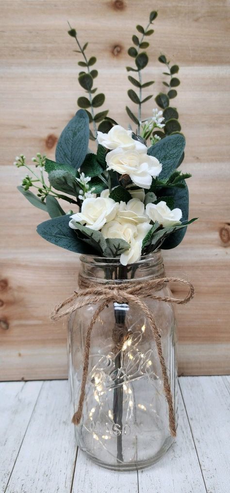 These beautiful faux mini-rose quart size mason jar centerpieces are a Glassy Gal original design. Each centerpiece includes one set of warm white LED fairy lights, a jute bow, a deluxe faux greenery arrangement with mini roses. You can choose your mini rose color at checkout.  These centerpieces are very popular for weddings, baby showers, parties, corporate events, holiday events, and any other event that requires table centerpieces. They also look great in any room of your house or office. To Boho Mason Jar Wedding Centerpieces, Mason Jar Centerpieces Table Decorations, Simple Centerpieces Wedding, Centerpieces Without Flowers, Jar Centerpiece Wedding, Barn Wedding Centerpieces, Mason Jar Flower Arrangements, Wedding Ceremony Readings, Wedding Centerpieces Mason Jars