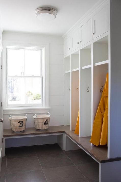 A nickel and white glass flush mount illuminates an l-shaped mudroom fitted with white shiplap walls and a wood top bench positioned beneath a window and continuing around to an adjacent wall fitted with white built-in open lockers fixed beneath white overhead cabinets accented with polished nickel knobs. Dream Mudroom, Industrial Baskets, White Shiplap Walls, Shiplap Trim, Black Shiplap, Transitional Laundry Room, Mudroom Remodel, Mudroom Cabinets, Shoe Organization