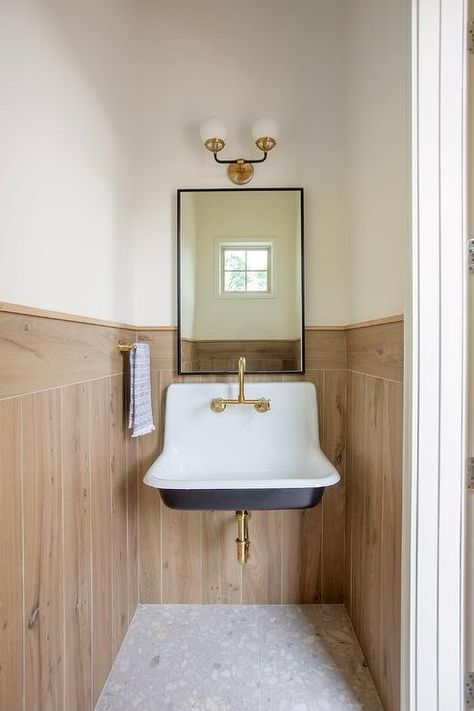 Welcoming powder room boasts a black and white vintage sink mounted against plank trim over gray recycled floor and beneath a black mirror lit by a black and gold sconce. Gold Mirror And Black Faucet, Powder Room With Pedestal Sink, Black Mirror Gold Faucet, Vintage Yellow And Black Tile Bathroom, Moody Powder Room Pedestal Sink, Bathroom With Pedestal Sink, Pedestal Sink Vintage, Pedistal Sink, Marble Tulip Table