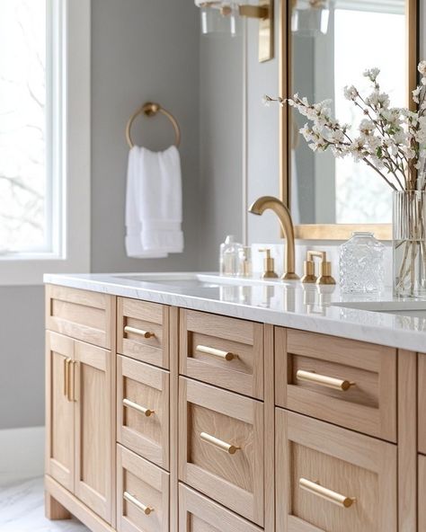 When it comes to creating a space that feels luxurious yet grounded, I love mixing natural tones and textures with sleek, modern fixtures. The oak cabinetry paired with the marble countertops and gold accents here creates that perfect balance of warmth and elegance, don’t you think? I’ve been obsessed with how the natural wood grains pop against the soft grey walls. And who knew a simple touch like fresh florals could transform the entire room? ⁣ ⁣ I’ve found that bringing in natural material... Light Wood And Gold Bathroom, Natural Oak Bathroom Vanity, Wood Vanity Gold Hardware, White Oak Kitchen Cabinets Gold Hardware, Light Wood Bathroom Cabinets, Gold Hardware Bathroom, White Oak Bathroom Vanity, White Oak Kitchen Cabinets, Wood Bathroom Cabinets