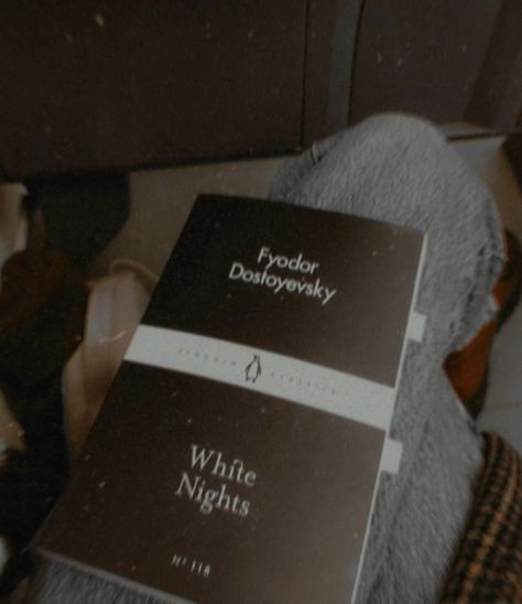 The photo is taken on a train, at a slight downward facing angle showing a close-up shot of a small pocket-book, resting on the photographer's jeans covered legs. The book is mostly a solid mat-black, with a white horizontal streak.

The image is quite dark and low-lit, monochrome in its colour palette, with slight shades of warm browns. The photo has a grainy overlay, giving the general appearance of being a very old photo. Perhaps from the late 20th to early 21st century. Russian Novels Aesthetic, White Nights Dostoevsky Book, Fyodor Dostoyevsky Books Aesthetic, Dark Obscure Literature, Fyodor Core Aesthetic, Fyodor Dostoyevsky White Nights, Fyodor Dostoyevsky Bsd Aesthetic, Old Literature Aesthetic, White Nights Dostoevsky Aesthetic