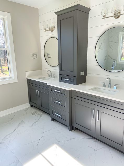 These beautiful slate grey master bathroom cabinets really pop against the nickle gap wall and marble floors. The round mirrors on either side of a center storage space ensure that two people have ample space and storage to get ready at the same time. Master Bath With Grey Cabinets, Bathroom Remodel Gray Cabinets, Dark Gray Bathroom Cabinets Master Bath, Marble And Shiplap Bathroom, Gray Floor And Cabinets, Grey Cabinets In Bathroom Master Bath, Grey Cabinets Master Bath, Farmhouse Bathroom Marble Floor, Master Bath Grey Floor