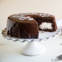 A square photo of the homemade ding dong cake taken from the side of the cake. There is a slice taken out of the cake and the creme filling is exposed. The white squiggle frosting is visible on the top of the cake, on top of chocolate frosting that drips down the side of the cake. The cake is on a white cake stand that has wave-like edges. It is taken against a white background. Homemade Ding Dongs, Ding Dong Cake Recipe, Chocolate Marshmallow Cake, Ding Dong Cake, Creme Filling, Marshmallow Filling, Hot Milk Cake, Kid Friendly Dessert, Square Photo