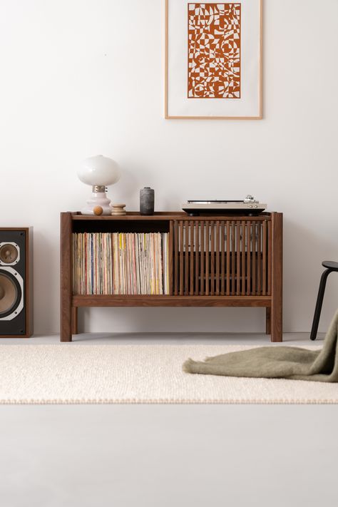 A mid-century modern styled room with a wooden sideboard holding vinyl records, a turntable, and decorative items including a white lamp and vases. Above the sideboard is a framed abstract artwork. To the left of the sideboard is a speaker, and to the right, part of a black chair is visible. The room has a white wall and a light-colored rug on the floor with a green blanket draped at the right edge. Small Tv Table Bohemian, Console Record Player Walmart, Cheap Record Player Stand Walmart, Record Player By Fireplace, Record Player Cabinet Black And Oak, Record Player Cabinet Target, Wood Record Player Table, Record Player Stand Coffee Tables, Record Player Table