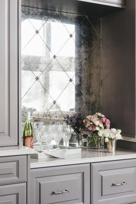 Gray kitchen bar features an antiqued mirror diamond pattern backsplash tiles positioned above a white quartz countertop framed by gray shaker cabinets fit with satin nickel hardware. Antique Mirror Backsplash Wet Bar, Mirror Tile Kitchen, Beveled Mirror Tile, Reflective Kitchen Backsplash, Mirrored Bar Backsplash, Antiqued Mirror Backsplash Kitchen, Smoked Mirror Tile, Aged Mirror Backsplash, Mercury Glass Tile Backsplash