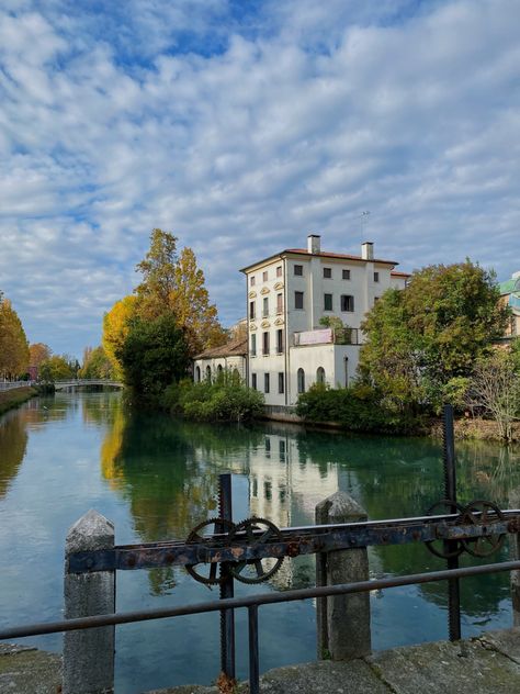 A white building sitting on the bank of a river in Treviso, Italy. The leaves are changing colors on the trees and the river is a beautiful blue. Italian City Aesthetic, Treviso Italy, Fake Insta, Italian City, Exchange Student, Thermal Bath, Italy Aesthetic, Northern Italy, Soft Summer