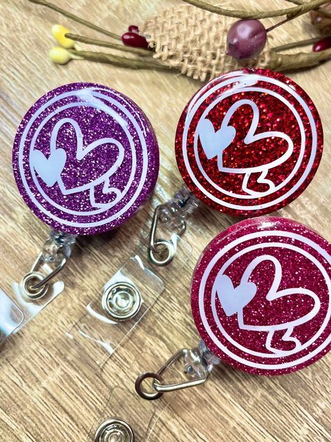 "Energize your work day with these fun custom made badge reels! Dimension: - Glitter Disk: 1.5\" wide - Reel Diameter: 1.25\" - Reel Cord length: Belt Clip 28\", Alligator Clip 23.6\" Available Clip Options: Belt Clip Swivel Alligator Clip Available Glitter Colors: Lilac Purple Mauve Ruby Red All will be made as shown in the picture. If you would like white vinyl instead of black, please note it in the personalization box and I can accommodate. Due to being a made to order, no returns are accept Backpack Hooks, Heart Glitter, Triple Heart, Badge Buddy, Office Staff, Tumbler Cups Diy, Colors Purple, Diy Resin Crafts, Nurse Badge Reel