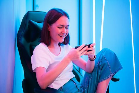 Cute gamer girl sit on a gaming chair, smiling and playing mobile online game on a smartphone. Having fun playing with friends online. Playing Video Game Poses Drawing, Playing With Friends, Gamer Chair, Chair Pose, Name For Instagram, Social Games, Mobile Gaming, Gaming Chairs, Sitting Poses