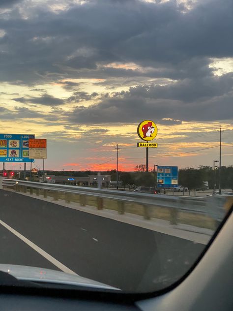 The picture is taken from the passenger seat of a car, looking out over a hot pink sunset far in the distance. In the foreground is a Highway sign listing local places to stop for food at the next exit (Taco Bell, McDonald’s, etc.) and a large sign for the gas station chain Buccee’s Bucees Texas Wallpaper, Texas Life Aesthetic, Bucees Texas Aesthetic, Buccees Aesthetic, Texas Astethic, Galveston Texas Aesthetic, Texas Summer Aesthetic, Atx Aesthetic, San Antonio Texas Aesthetic