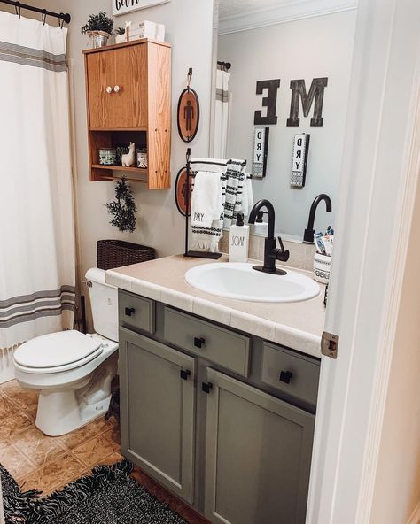 A Evergreen Fog by Sherwin Williams bathroom vanity with black knobs is topped by a beige countertop. The vanity stands under a beveled mirror mounted next to a stained wood over the toilet cabinet. The cabinet is fitted beside a white and gray shower curtain hanging from a black rod. Evergreen Fog Bathroom, Bathroom With Wallpaper, Sherwin Williams Evergreen Fog, Dark Grey Bedding, Gray Shower Curtain, Countertop Covers, Evergreen Fog, Toilet Cabinet, Black Metal Shelf