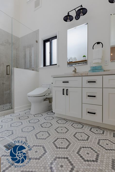 A white bathroom with black hardware and light grey mosaic tiles is seen. The ceilings have been vaulted and a dividing wall removed to open the space between the double vanity on the right and toilet with barrier free shower niche on the left. The cabinets are semi-custom, painted white, with black hardware. The countertop is quartz and features new his and hers sinks. Above each sink is a frameless mirror with a black double light fixture overhead. Toilet Between Shower And Vanity, Closed In Toilet Master Bath, Half Wall Between Shower And Toilet, Shower With Half Wall And Glass Door, Master Bath Seperate Toilet, Master Open To Bathroom, Custom Vanity Cabinets, His And Hers Sinks, Shower Wall Tile