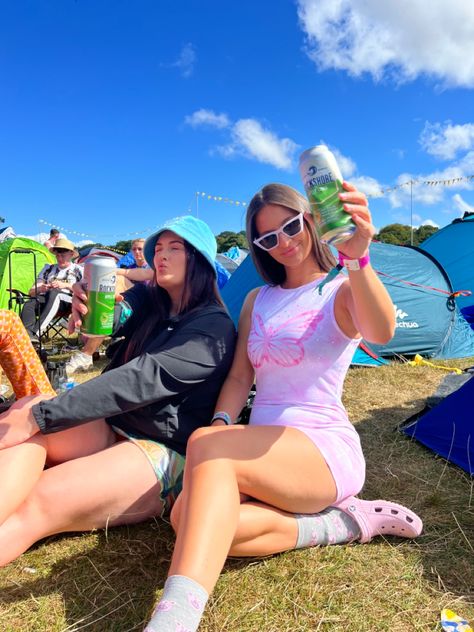 Two girls one in pink other in blue cheersing infront of tents whilst sitting on floor of campsite at a festival