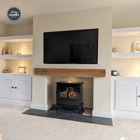 A neutral living room with a stunning large renovated fireplace. The walls are a cream colour and there is a slate hearth in front of the fireplace. There is a sleek, black log burner inside the fireplace and a long wooden mantel just above. The TV is wall mounted above the fireplace and fitted inside the wall. To either side of fireplace are alcoves with white floating shelves fitted. There are white cabinets under the shelves on each side and warm LED lights illuminating the shelves. Oak Beam Fireplace, Mantel Beam, Oak Mantle, Oak Mantel, Loft Style Bedroom, Oak Fireplace, Real Wood Furniture, Feature Wall Living Room, Wooden Mantel