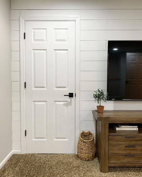 A white 6-panel door with black hardware sits on the side of a white shiplap living room wall. A black flatscreen TV hangs near a plant in an orange clay pot on a wooden side table. A wicker basket is placed on the beige carpet. 6 Panel Door With Black Hardware, White 6 Panel Interior Doors, Black Hardware On Doors, Interior Doors With Black Hardware, White Doors Black Hardware, White Door Black Hardware, White Doors With Black Hardware, White Shiplap Living Room, Doors With Black Hardware