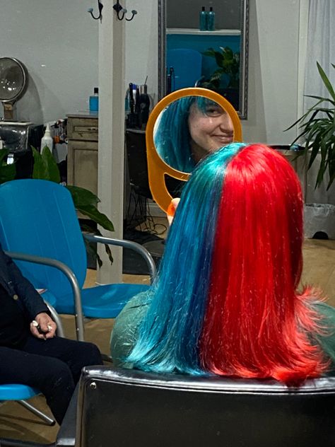 A view of a girl turned away from the camera sitting in a chair at the hair salon. Her hair is split half blue and half red and she is holding a mirror where you can see a piece of her face smiling. Blue And Red Split Dye, Half Red Half Blue Hair, Red And Blue Hair Split, Red And Teal Hair, Red Split Dye, Blue Split Dye, Red And Blue Hair, Split Dye Hair Ideas, Blue And Red Hair