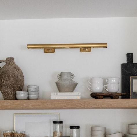 Visual Comfort & Co. on Instagram: "Shelfie appreciation post. 😍 Designer @rachaelbialkowski of @thelifestyledco⁣⁣⁣⁣⁠ helped her client @suzannavilla create a fresh yet timeless kitchen using a mix of neutral millwork and organic elements. Selected by the homeowner, the "cookies and cream" backsplash and countertop make a bold statement while pairing gorgeously with the white oak shelves for a natural look. Overhead, our iconic 30" Cabinet Maker's Picture Light by Chapman & Myers adds classic appeal to the space in a radiant Hand-Rubbed Antique Brass finish. The fixture brings a soft glow to the thoughtful collection of entertaining necessities and artful objects below. ​Architect: @harrisondesign 📷: @elizabethlaurengranger" Gallery Kitchen Lighting Ideas, Picture Light In Kitchen, White Oak Shelves, Cream Backsplash, Kitchen Built Ins, Kitchen Bookshelf, Kitchen 2023, Bookshelf Lighting, Lamp Kitchen