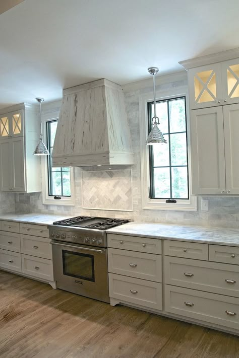 Grey kitchen. Stunning transitional kitchen is equipped with a rustic pecky cypress hood fixed between windows framed by gray marble backsplash tiles illuminated by industrial pendants. The hood is located above herringbone marble cooktop tiles accenting a Thermador range flanked by white drawers with polished nickel cup pulls topped with gray and white marble countertops positioned beneath white upper cabinets finished with x front lighted display cabinets. According to the designer, the ca... Kitchen Cabinets Grey And White, Kitchen Window Design, Kitchen Cabinet Trends, Серая Кухня, Hood Design, Kitchen Hood, White Shaker Cabinets, Herringbone Backsplash, Classic Kitchen