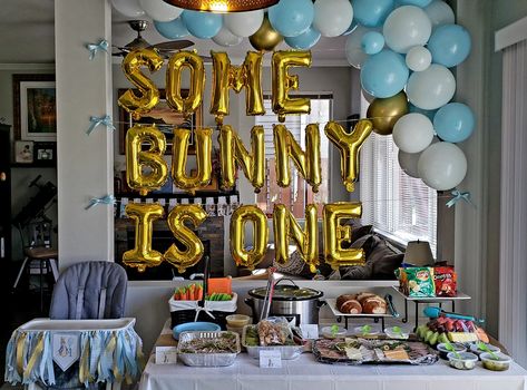 Picture shows a table with food, balloon arch over table with blue, gold, and white balloons. In between the table and baloon arch are balloon letters that say, "some bunny is one". There is a high chair located beside the table with a Peter rabbit tag and blue ribbon. Easter First Birthday Boy, Somebunny Is Turning One Boy, Some Bunny Is One Birthday Boy, Some Bunny Is Turning One Boy, Peter Rabbit Theme Party, One Year Old Birthday Party, Peter Rabbit Theme, One Year Old Birthday, Easter Birthday Party