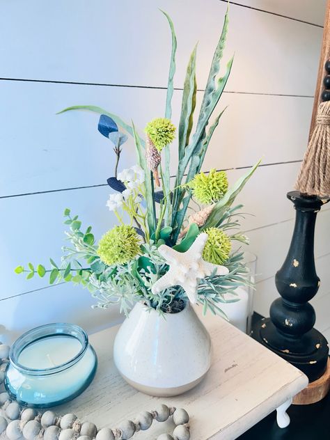 This bright and refreshing small coastal flower arrangement in a ceramic small planter is beautiful! This arrangement is made of mixed green faux greenery and accented with seashells and starfish.  **Display stand and styling items are not included in the purchase** THIS IS A SMALL ARRANGEMENT DIMENSIONS BELOW:  This arrangement is 14 inches high, 6 inches wide.  Ceramic  planter is 4x4 inches.  Greenery is permanently attached inside of the planter.  All sides of the centerpiece are displayable.  Suggested indoor or covered outdoor use.  Please message for custom orders! Ocean Theme Flower Arrangements, Nautical Flower Arrangements, Coastal Flower Arrangements, Beach Flower Arrangements, Coastal Arrangements, Coastal Florals, Pink Orchids Centerpiece, Beachy Centerpieces, Beach Theme Centerpieces