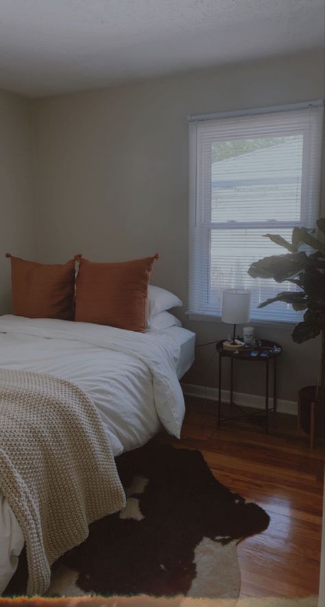 white bed with a taupe knit blanket and two large orange throw pillows, underneath is a brown abstract shape cowhide rug, and next to the bed is a black metal nightstand and a fiddle fig plant Cow Rug Bedroom Decor, Cowhide Rug Bedroom Ideas, Bedroom With Cowhide Rug, Cow Hide Rug Bedroom, Cow Hide Rug Bedroom Ideas, Ikea Cow Rug, Cow Rug Bedroom, Cowhide Bedroom Ideas, Room Ideas Western