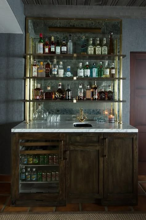 Chic rustic wet bar boasts dark stained oak cabinets fitted with a glass door beverage fridge topped with white marble lined with a bar sink and an antique brass vintage faucet under stacked wood and brass shelves lining an antiqued mirrored backsplash. Antique Mirror Backsplash, Vintage Faucet, Glass Shelves Kitchen, Floating Glass Shelves, Brass Shelves, Bar Shelves, Mirror Backsplash, Bar Designs, Rustic Bar