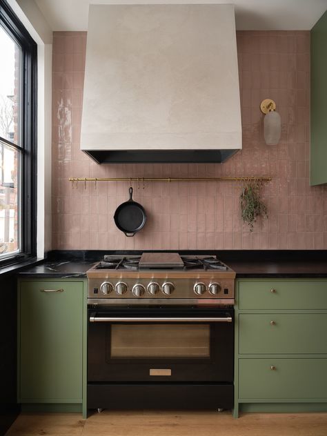 A beautiful green kitchen with tiles on the backsplash that are a taupe pink colour that pairs really well with the medium green of this kitchen. Along with all the brass accent with the handles, the rail, the sconce, the overall material palette of this project gives off a very unique and english vibe. The project is vibrant and colourful. We see big black windows that matches the dark soapstone countertop. Pink Kitchen Walls, Green Kitchen Inspiration, Sage Kitchen, Pink Cabinets, Green Kitchen Designs, Green Backsplash, Colourful Kitchen, Sage Green Kitchen, Green Kitchen Cabinets