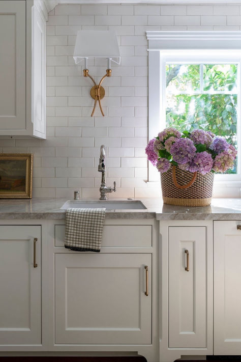 To leave the center island unobstructed, designer @heatherlunddesign added a charming prep sink to the perimeter counter of this timeless kitchen. Perfect for chopping produce or trimming flowers from the garden, this versatile space is accented with classic forms, mixed metal accents, and neutral tones. Above the gray quartzite and traditional cabinetry, our Westerly Double Sconce by Chapman & Myers elevates the off-white tile backsplash. Stylist: @tcbstyle 📷: @jessicabrydsonphotography Light Over Kitchen Sink, Over Kitchen Sink, Classic Kitchen Design, White Tile Backsplash, Neutral Kitchen, Prep Sink, Classic Lighting, Timeless Kitchen, Traditional Cabinets