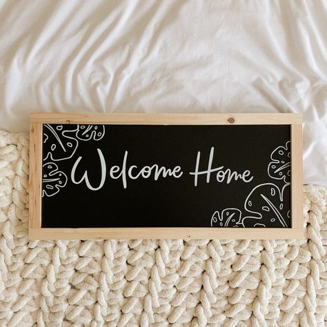 A black chalkboard with ‘welcome home’ written in white pen and white leaves drawn around the edges Welcome To Chalkboard Sign, Welcome Home Chalk Art, Welcome On Black Board, Chalk Welcome Sign, Home Sweet Home Chalkboard Art, Welcome Home Signs, Black Chalkboard, Beautiful Lettering, Welcome Home